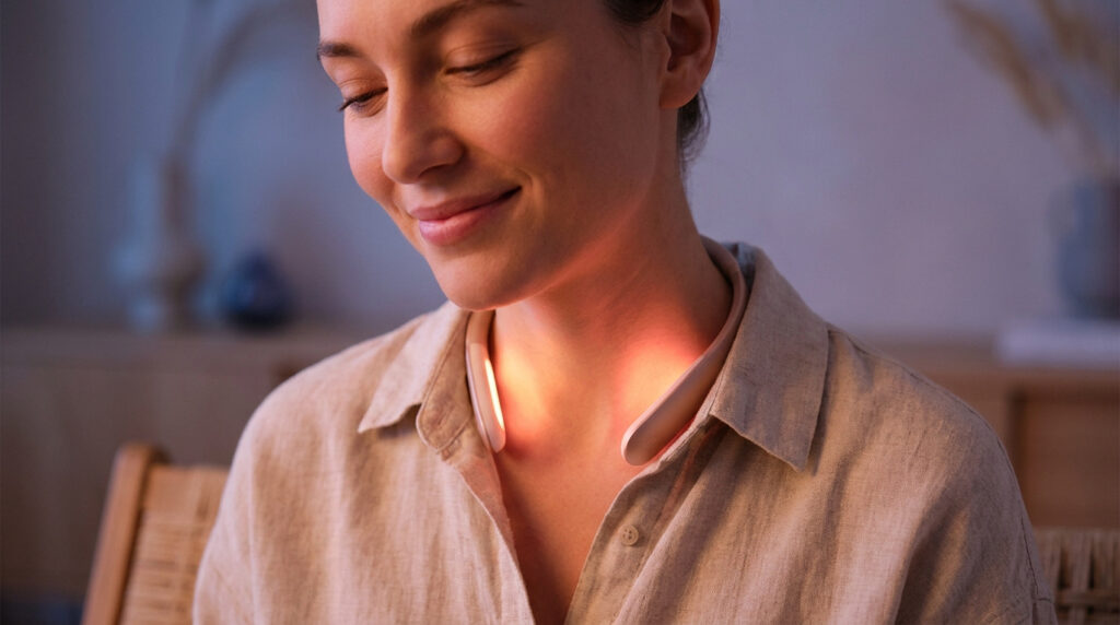 Une femme souriante porte un appareil de luxothérapie infrarouge autour du cou dans un intérieur chaleureux.